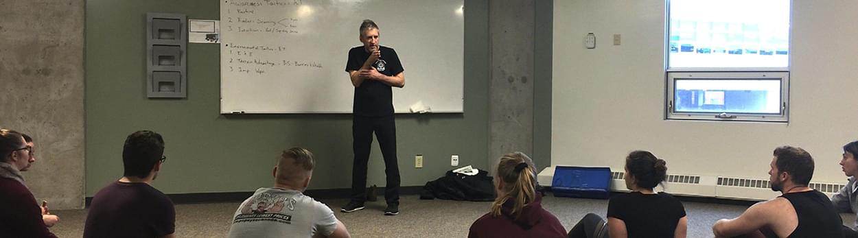 Self defense coach Calen Paine stands in front of white board teaching group of seminar students, seminars for businesses and organizations, Edmonton